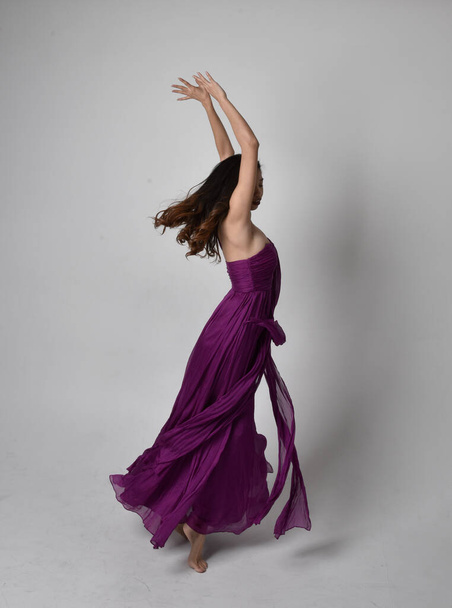 紫の流れるガウンを着てかなりブルネットのアジアの女の子の完全な長さの肖像画。立ち、スタジオで踊るポーズ. - 写真・画像