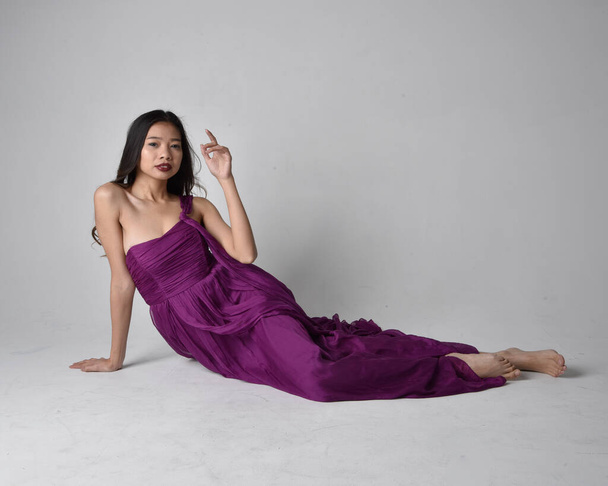 Полный портрет симпатичной брюнетки азиатской девушки в фиолетовом плаще. Сидя на студийном фоне. - Фото, изображение