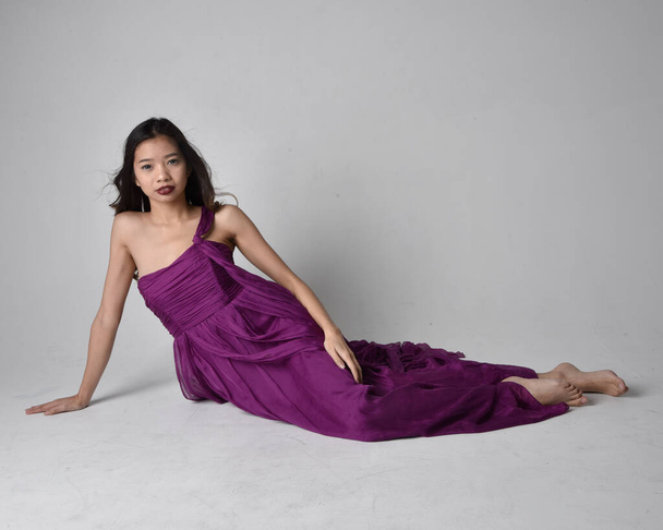 Full length  portrait of pretty brunette asian girl wearing purple flowing  gown. Sitting pose on on studio background. - Photo, Image