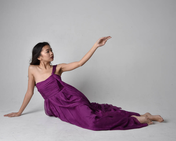 Full length  portrait of pretty brunette asian girl wearing purple flowing  gown. Sitting pose on on studio background. - Fotó, kép