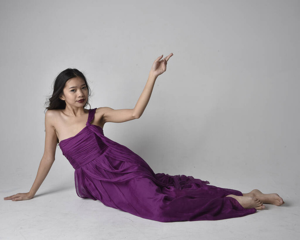 Full length  portrait of pretty brunette asian girl wearing purple flowing  gown. Sitting pose on on studio background. - Photo, image