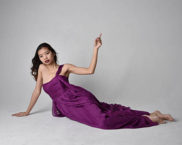Full length  portrait of pretty brunette asian girl wearing purple flowing  gown. Sitting pose on on studio background. - Foto, immagini