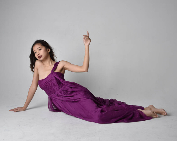 Full length  portrait of pretty brunette asian girl wearing purple flowing  gown. Sitting pose on on studio background. - 写真・画像