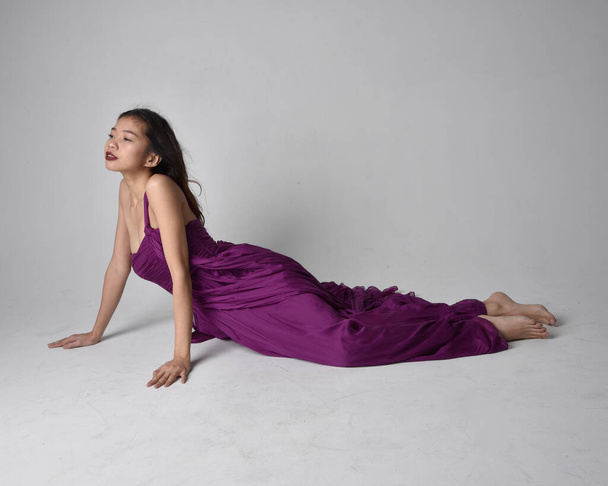 Full length  portrait of pretty brunette asian girl wearing purple flowing  gown. Sitting pose on on studio background. - Fotó, kép