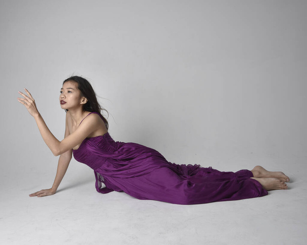 Full length  portrait of pretty brunette asian girl wearing purple flowing  gown. Sitting pose on on studio background. - Foto, Imagem