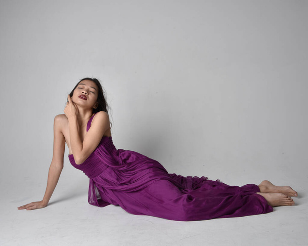 紫の流れるガウンを着てかなりブルネットのアジアの女の子の完全な長さの肖像画。座席ポーズ上のスタジオ背景. - 写真・画像