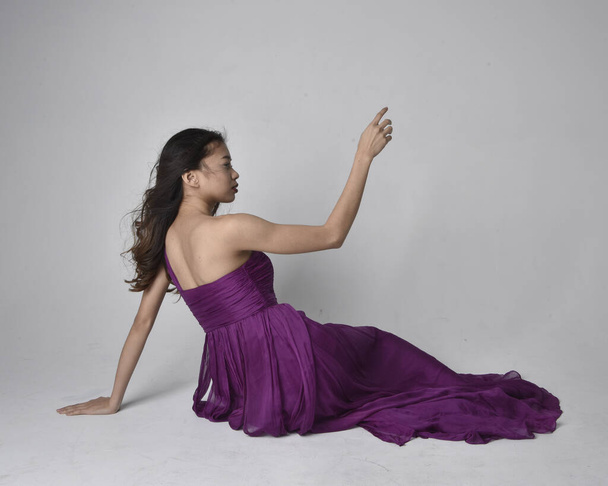 Полный портрет симпатичной брюнетки азиатской девушки в фиолетовом плаще. Сидя на студийном фоне. - Фото, изображение