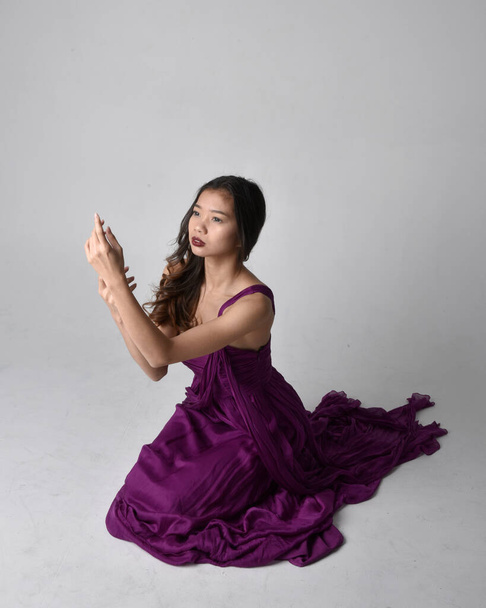 Full length  portrait of pretty brunette asian girl wearing purple flowing  gown. Sitting pose on on studio background. - Zdjęcie, obraz