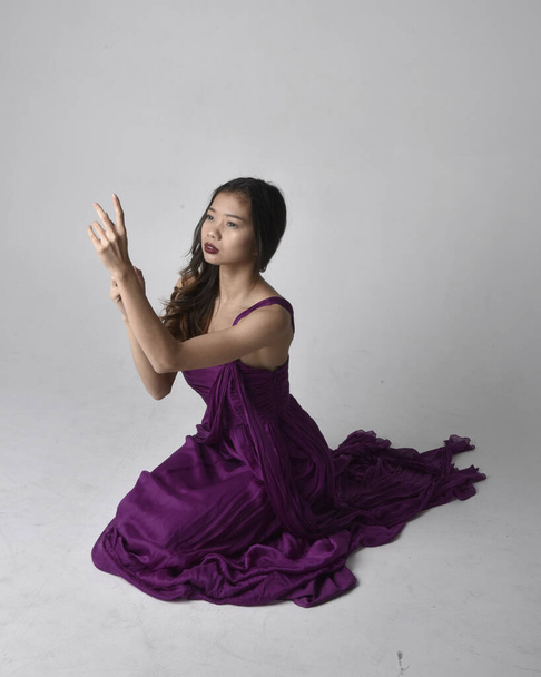 Ganzes Porträt der hübschen brünetten asiatischen Mädchen trägt lila fließenden Kleid. Sitzpositionen auf Studiohintergrund. - Foto, Bild