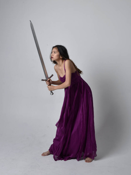 Full length  portrait of pretty brunette asian girl wearing purple flowing  gown. Standing pose holding a sword  on on studio background. - Foto, imagen