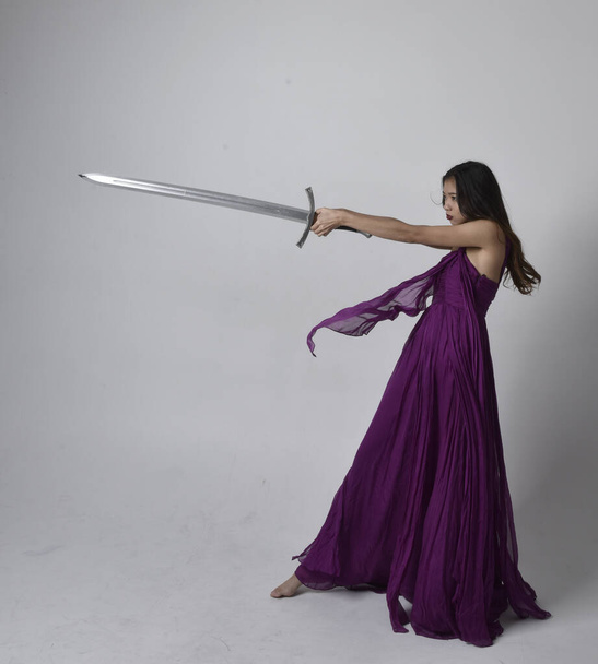 Full length  portrait of pretty brunette asian girl wearing purple flowing  gown. Standing pose holding a sword  on on studio background. - Foto, imagen