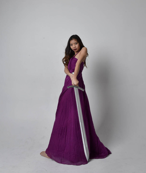 Ganzes Porträt der hübschen brünetten asiatischen Mädchen trägt lila fließenden Kleid. Stehende Pose mit einem Schwert auf Studiohintergrund. - Foto, Bild