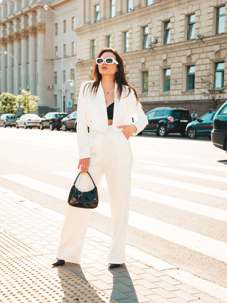 Portrait of young sexy modern businesswoman model. Hot beautiful woman in white suit posing on the street background. Fashion female walking outdoors in sunglasses - Fotó, kép