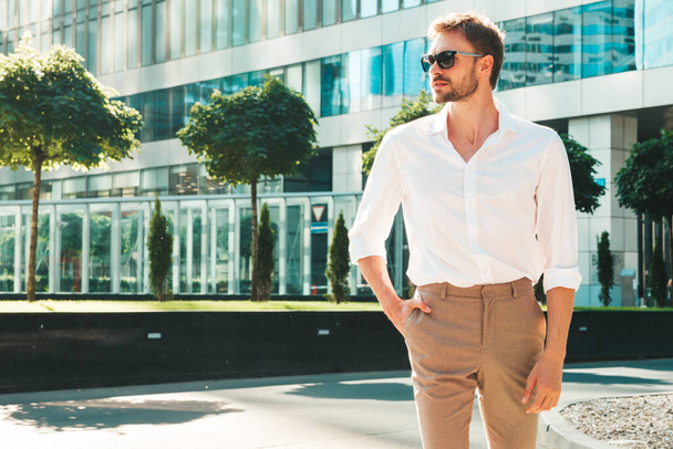 Portrait of handsome smiling stylish hipster lambersexual model.Modern man dressed in white shirt. Fashion male posing in the street background near skyscrapers in sunglasses - Fotografie, Obrázek
