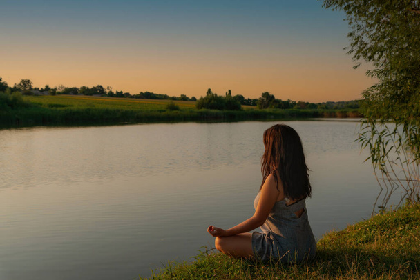 Woman meditating in lotus pose during sunset. Girl meditates on shore of river alone with herself. Yoga, meditation, tranquility, relax in nature. Beatiful landscape. - Zdjęcie, obraz