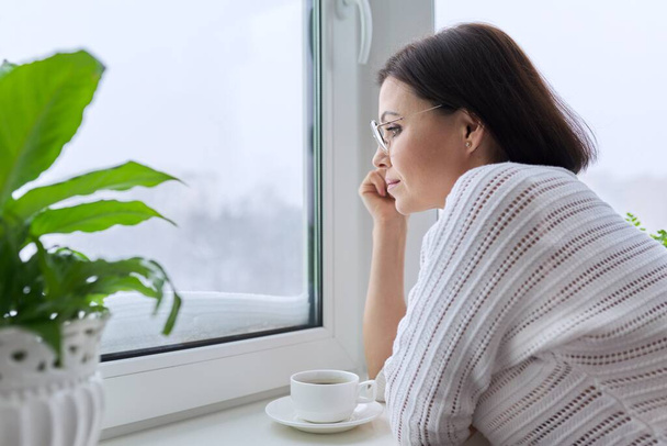 Sezóna zima, zasněžený den, usmívající se žena středního věku s šálkem kávy dívající se z okna. - Fotografie, Obrázek