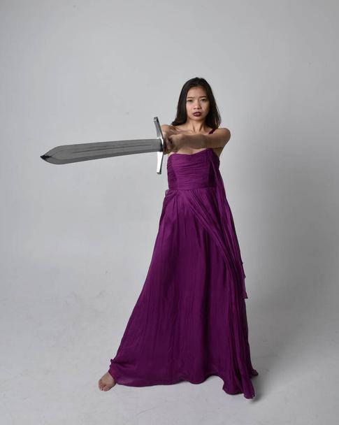Full length portret van mooie brunette aziatische meisje dragen paars vloeiende jurk. Staande houding met een zwaard op studio achtergrond. - Foto, afbeelding