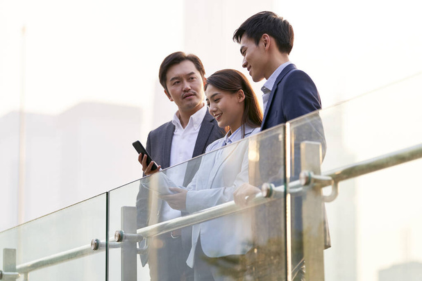 tres feliz asiático empresarios buscando en celular charlando hablando de negocios al aire libre - Foto, Imagen