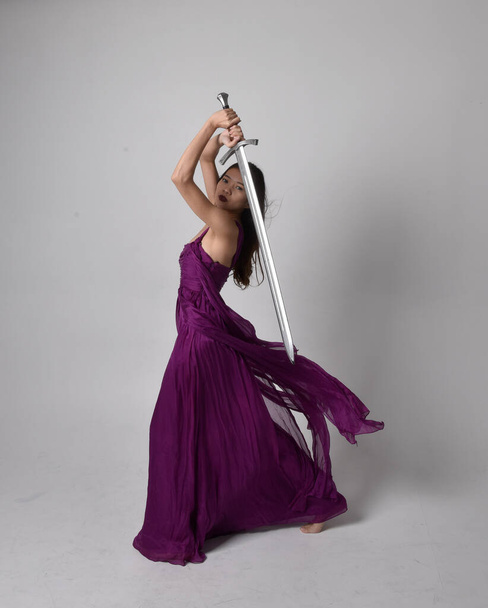 Full length  portrait of pretty brunette asian girl wearing purple flowing  gown. Standing pose holding a sword  on on studio background. - Zdjęcie, obraz