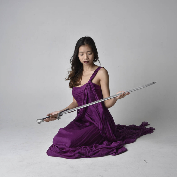 Ganzes Porträt der hübschen brünetten asiatischen Mädchen trägt lila fließenden Kleid. Sitzende Pose mit einem Schwert auf Studiohintergrund. - Foto, Bild