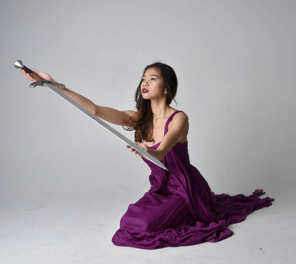 Full length  portrait of pretty brunette asian girl wearing purple flowing  gown. Sitting pose holding a sword  on on studio background. - Foto, immagini