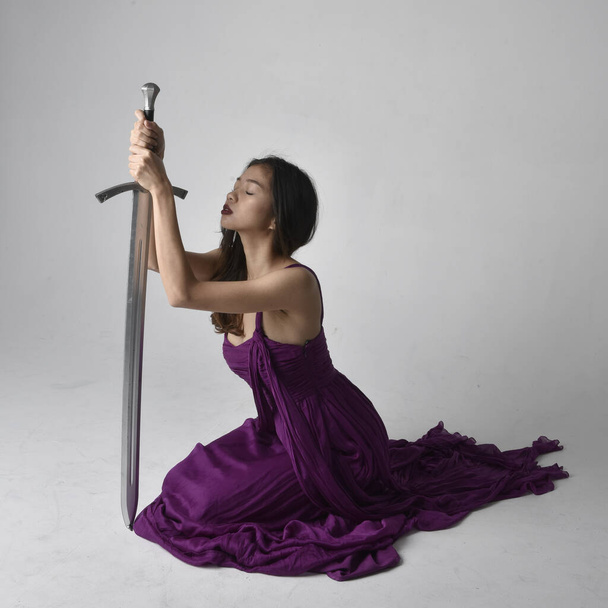 Ganzes Porträt der hübschen brünetten asiatischen Mädchen trägt lila fließenden Kleid. Sitzende Pose mit einem Schwert auf Studiohintergrund. - Foto, Bild