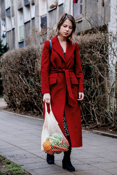 Stilvolle Frau in rotem Mantel und Netztasche, die nach dem Einkaufen nach Hause wacht. Osteuropa  - Foto, Bild