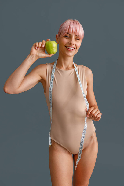 Усміхнена струнка молода жінка в бежевому костюмі тримає зелене яблуко і вимірювальну стрічку, стоїть ізольовано на сірому студійному фоні
 - Фото, зображення