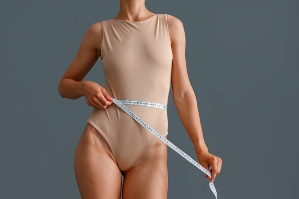 Cropped shot of slim young woman in beige bodysuit measuring her waist with a measuring tape, standing isolated over gray studio background - Photo, Image