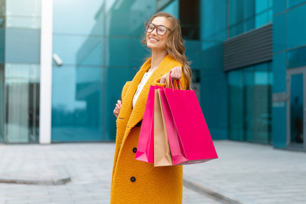 Business woman with shopping bags dressed yellow coat walking outdoors corporative building background Caucasian female business person near office building Stylish businesswoman season sale - Fotoğraf, Görsel