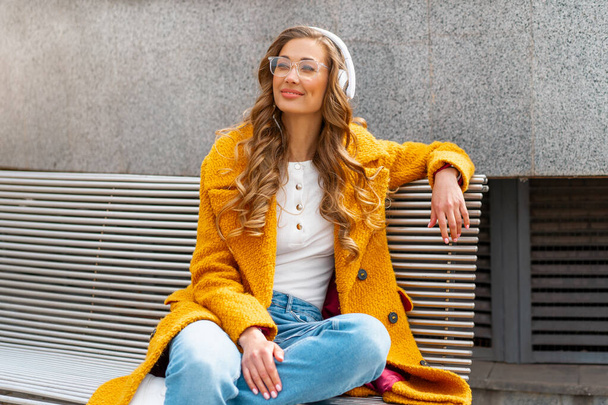 Woman Eyeglasses Listen Music Headphone Outdoor Sitting Bench Outdoor Dressed Stylish Yellow Coat Smile Caucasian Female 30s Enjoy Podcast Or Audio Books Outside  - Foto, afbeelding