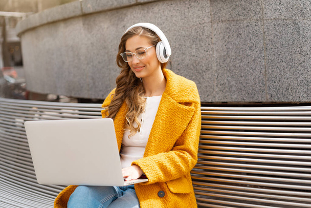 Business Woman Wear Eyeglasses Listen Music Headphone Outdoor Sitting Bench Using Laptop Outdoor Dressed Stylish Yellow Coat Smile Caucasian Female 30s Enjoy Podcast Or Audio Books Outside  - Foto, immagini