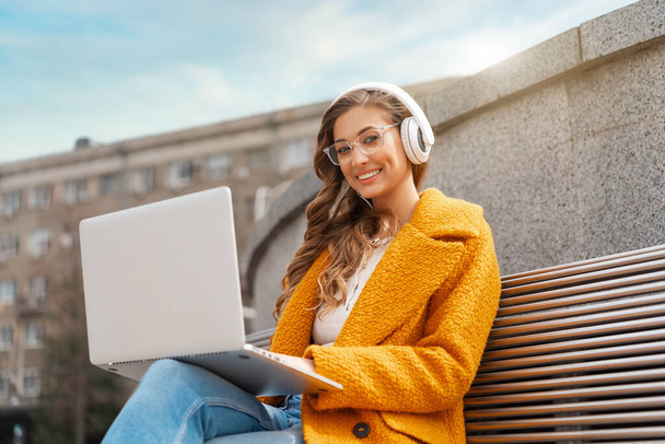 Business Woman Wear Eyeglasses Listen Music Headphone Outdoor Sitting Bench Using Laptop Outdoor Dressed Stylish Yellow Coat Smile Caucasian Female 30s Enjoy Podcast Or Audio Books Outside  - Foto, afbeelding