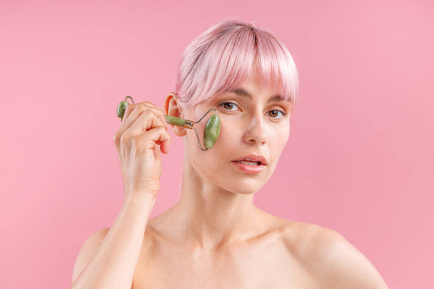 Portrait de jeune femme nue aux cheveux roses utilisant un rouleau facial en jade naturel pour les soins de la peau, posant isolé sur fond rose - Photo, image