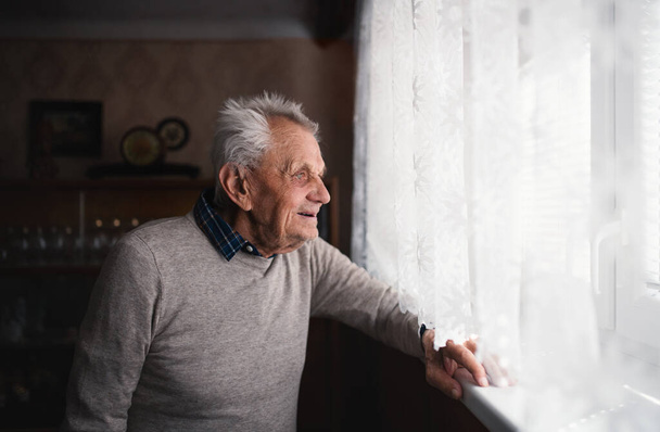 Portrait of elderly man standing indoors at home, looking out through window. - 写真・画像