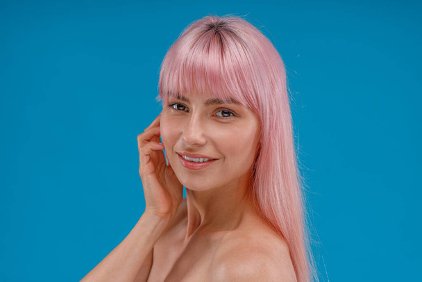 Πορτρέτο της όμορφης νεαρής γυναίκας με τα ροζ μαλλιά και το τέλειο δέρμα χαμογελώντας στην κάμερα, θέτοντας απομονωμένη πάνω από το μπλε φόντο στούντιο - Φωτογραφία, εικόνα