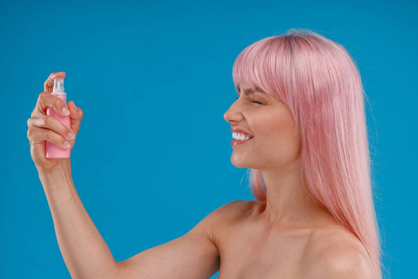 Portrait of cheerful woman with pink hair using spray in plastic bottle over her face with closed eyes, posing isolated over blue studio background - Fotografie, Obrázek