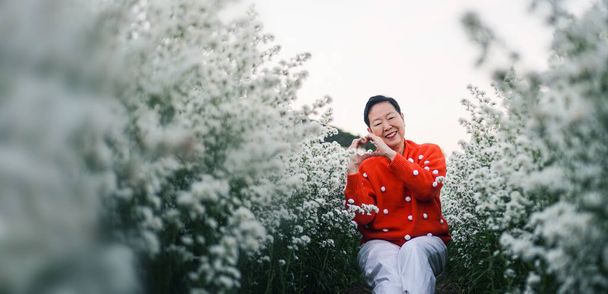 Asian senior elderly woman smiling celebrating Christmas holiday season in white flower field with red sweater - Photo, image