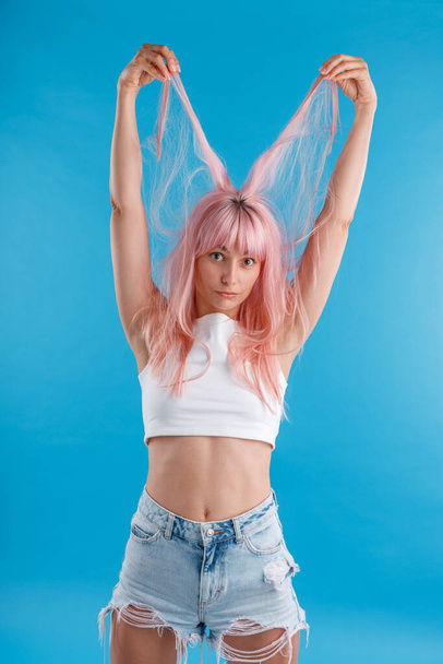 Забавная женщина-модель в повседневной одежде играет с розовыми волосами, держа нити над головой, стоя изолированно на синем фоне студии - Фото, изображение