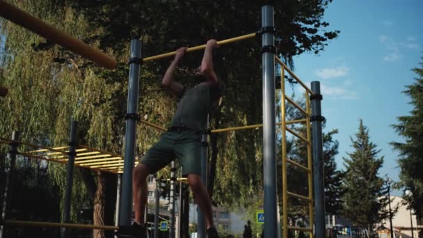 Sports activities on outdoor ground and healthy lifestyle. Fitness and strength exercises for muscles of arms. Muscular Caucasian man with beard pulls himself up on horizontal bar with two hands. - Footage, Video