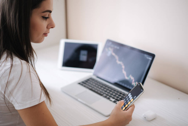 Atraktivní mladá žena sedí u stolu před notebookem, tablet a používá chytrý telefon pro práci. Krásná brunetka dívka analyzuje akciový trh, zatímco doma - Fotografie, Obrázek