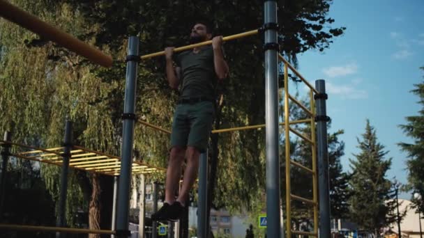 Sports activities on outdoor ground and healthy lifestyle. Fitness and strength exercises for muscles of arms. Muscular Caucasian man with beard pulls himself up on horizontal bar with two hands. - Footage, Video