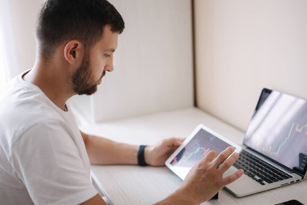 Hombre mirando gráfico de finanzas en el ordenador portátil y tableta, comerciante día, invertir desde casa. El joven supervisa sus inversiones. Hombre de negocios ambicioso - Foto, imagen