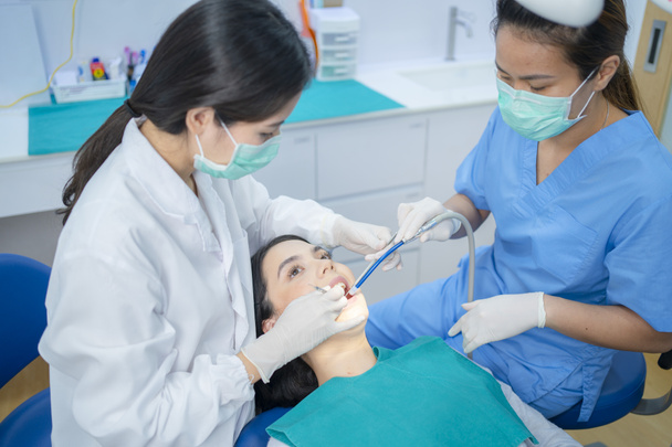 フェイスマスクを着用しているアジアの若い歯科医は、口腔ケア治療のための医療機器を使用します。吸引機を使用して横に座っているアシスタントは、クリニックでの治療中に患者の口を乾燥させます. - 写真・画像