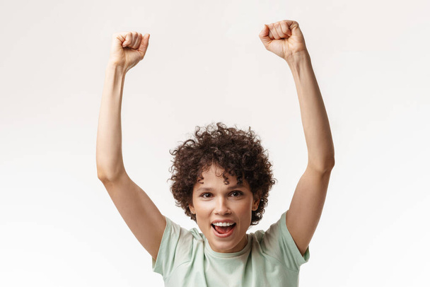 Young curly excited woman screaming while winner gesture isolated over white background - Фото, изображение