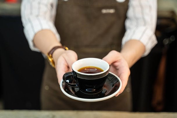 Close up of Unrecognised Waitress stand and handing a cup of hot coffee to customer with two hands at cafe. Business owner girl barista in apron work in restaurant serving occupation service to people - Foto, Imagem