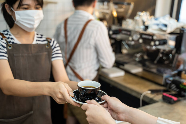 Close up of Unrecognised Waitress stand and handing a cup of hot coffee to customer with two hands at cafe. Business owner girl barista in apron work in restaurant serving occupation service to people - Photo, Image
