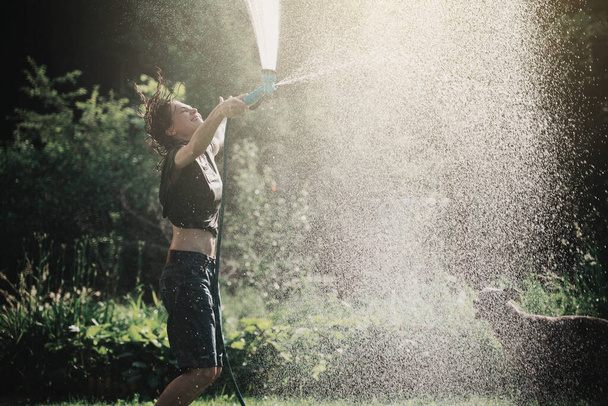 Beautiful young cheerful woman pours herself with water from a sprinkler in a green garden on a hot summer day - Φωτογραφία, εικόνα