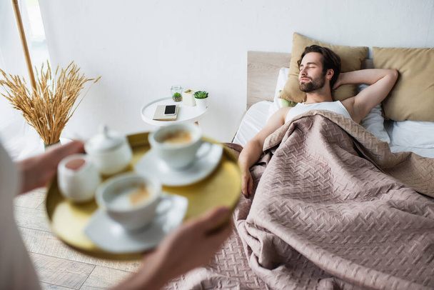 blurred woman holding tray with cups of coffee near sleeping man  - Foto, Imagen