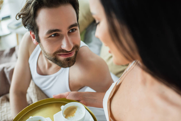 blurred woman holding tray with cups of coffee near bearded boyfriend in bed - Fotó, kép